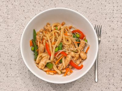 A bowl of udon noodles with sliced chicken, snap peas, red pepper, carrots and edamame