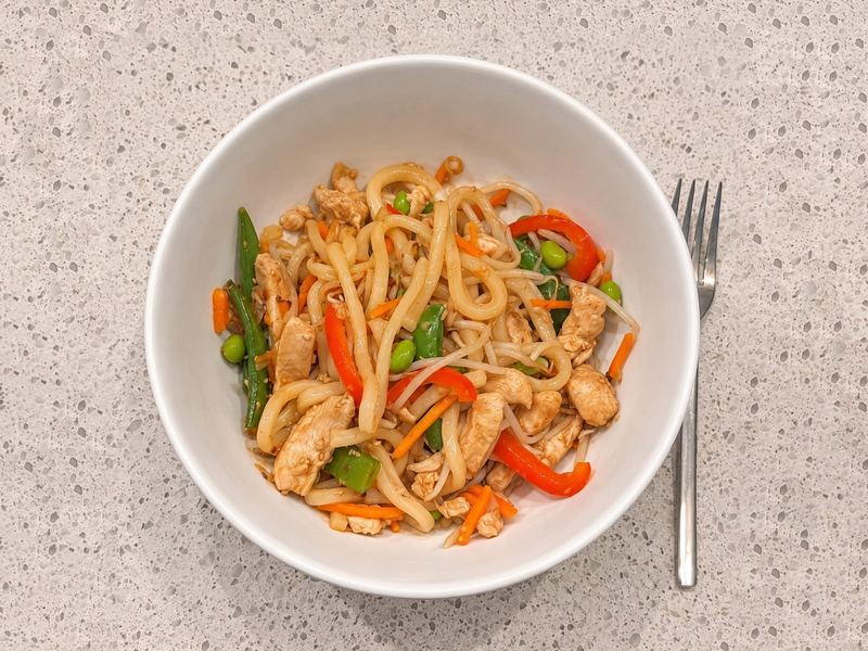 A bowl of udon noodles with sliced chicken, snap peas, red pepper, carrots and edamame
